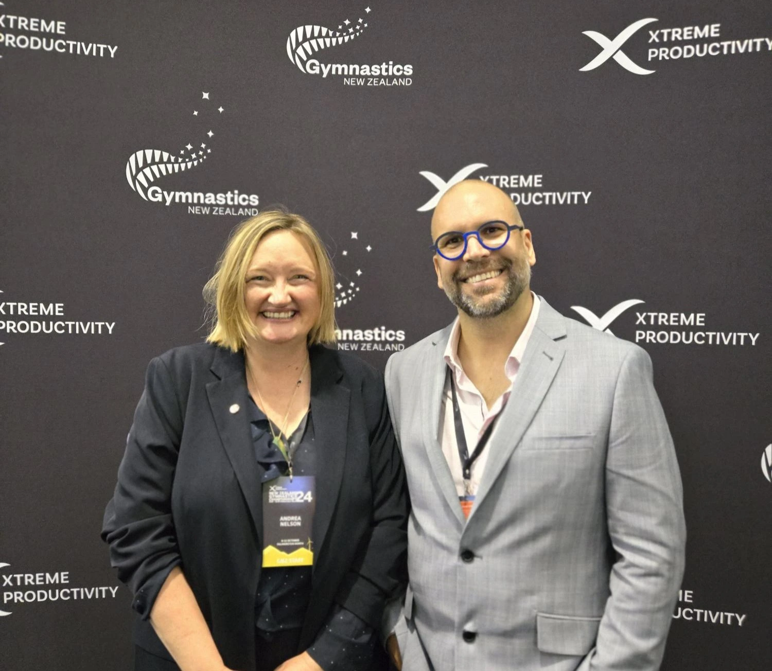 Two individuals, Christi Moller and Andrea Nelson standing together in front of a black backdrop at the XP Gymnastics Championships 2024, smiling for the camera